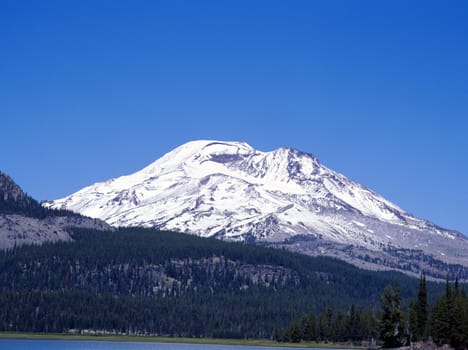 Snow covered mountain in the Cascades range next to a clear lake and pine tree forest