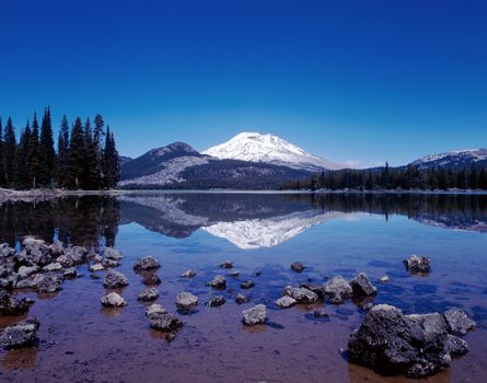 Mountain in Cascades range in Oregon reflected in a lake with clear blue sky and pine trees on either side