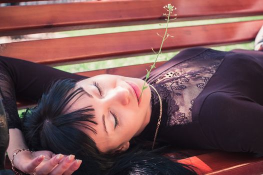 Young woman enjoying the sun, lying on a bench, with closed eyes and a flower in her lips