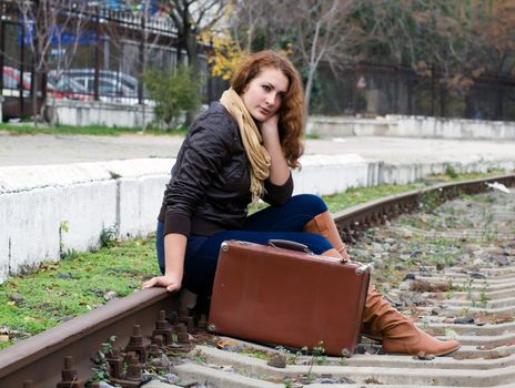 Beautiful girl sitting on a suitcase along the train tracks