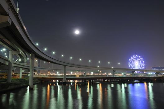  hanged-up highway road over Tokyo bay waters at night time with moon and ferris-wheel illumination on backward
