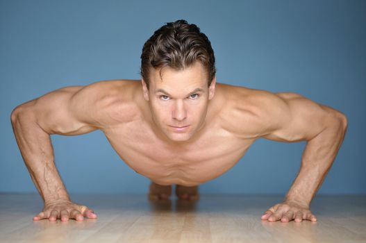 Handsome muscular man looks at camera while performing pushup on floor with blue wall background