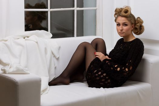 The girl with the avant-garde hairstyle in black sitting on a white sofa against the window