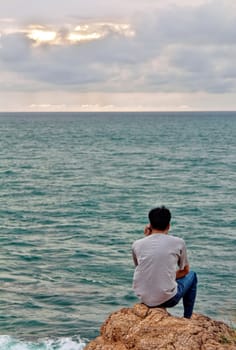 A man looks into the sea while sitting on a rock in the background of a cloudy sunset sky