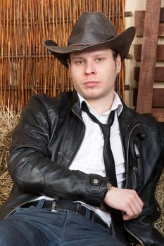 Glam cowboy in leather jacket in the rural interior is sitting on a haystack.