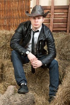 A young cowboy in leather jacket in the rural interior is sitting on a haystack.