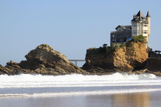 An old mansion perched on a rock in the ocean near a beach on a blue sky background