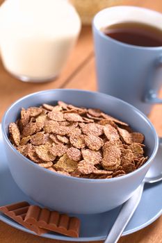 Bowl of chocolate corn flakes cereal with spoon and chocolate bars, a cup of tea and a jug of milk in the back (Selective Focus, Focus in the middle of the cereal)