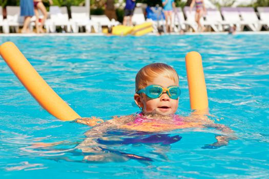 Little girl wearing swimming goggles learning to swim with pool noodle
