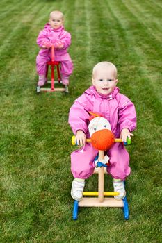 Two little baby girls wearing pink suits sitting on rocking horses outdoors