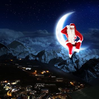 photo of santa claus sitting on the moon with a city and mountains below