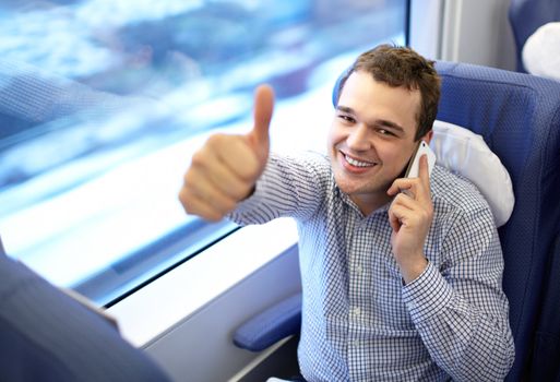 Young successful smiling businessman in modern high speed train is talking smartphone and showing his thumb up