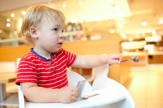 Little boy is sitting in the baby chair and holds the spoon in the restaurant.