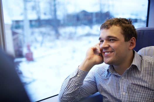 The young man is sitting by the window in the modern high-speed train and  is talking on the phone and smiling.