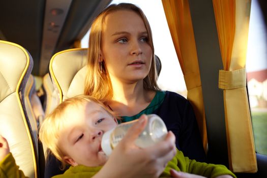 Young mother is feeding her two year old son with milk, while they are travelling by intercity bus. Close up portraits.
