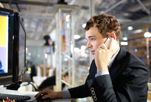 The young man works at a computer and talking on the phone.