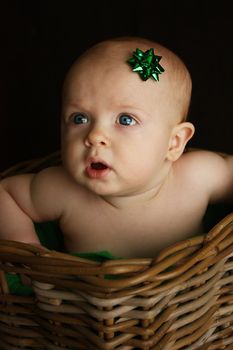 A close up of an infant with a green Christmas gift bow on his head, sitting in a wicker basket,  on a black background.