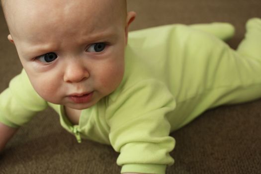 A close-up of a baby in a light green outfit on a brown carpet, crying.