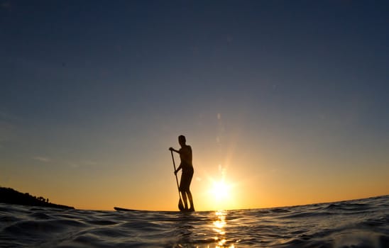 A young man paddles his surfboard in to shore at sunset
