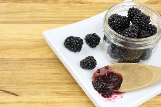 Blackberries in a jar, with blackberry jam on a wooden spoon, all on a white plate sitting on a wooden countertop.