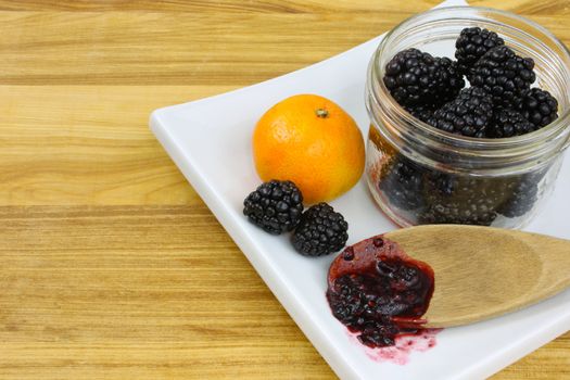 Blackberries in a jar, with a clementine orange and a wooden spoon of blackberry jam on a white plate sitting on a wooden countertop.







Three jars of home-made blackberry jam and three blackberries on a wooden countertop.