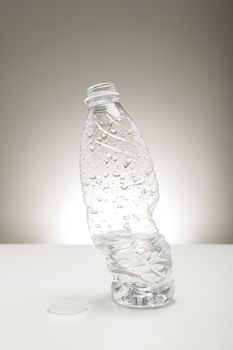 Twisted distorted clear plastic bottle of water covered in beaded water droplets standing upright on a studio countertop on a neutral grey background with highlight