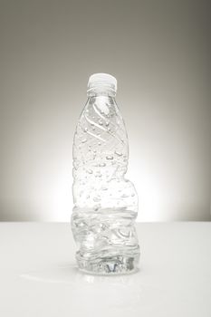 Half consumed bottle of water in a clear transparent bottle that has been partially twisted in readiness for recycling standing on a studio countertop on a grey background with highlight