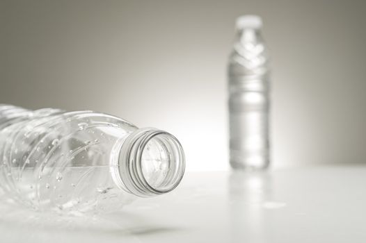 Closeup cropped view of the neck of an empty plastic water bottle lying on its side with a full bottle of water visible in the distance on a neutral grey background