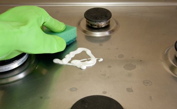 Hand in protective green glove is polishing a gas stove with sponge