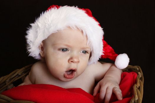 A close-up of a baby in a basket, wearing a santa hat, with an open-mouthed scolding expression, or expression of shock, isolated on a black background.