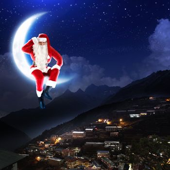 photo of santa claus sitting on the moon with a city and mountains below