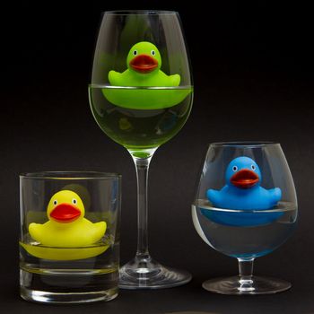 Green, yellow and blue rubber duck in different glasses on a dark background