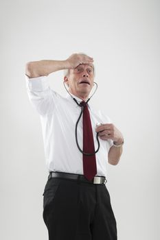 Fun portrait of a hypochondriac man worried by his health with one hand to his forehead while listening to his heart using a stethoscope with the other, or as a warning of heart disease in the elderly