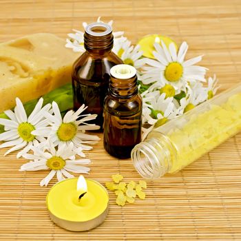 Oil in two different bottles, two homemade soap, bath salt, two candles with flames and chamomile flowers on a bamboo mat