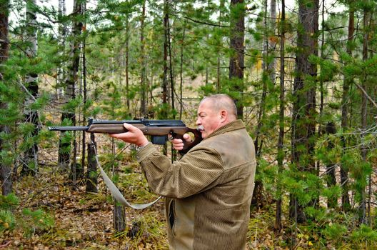 An older man in a brown jacket with a rifle in his hands on a background of trees