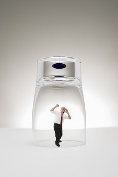 Conceptual studio image of a diminutive grey-haired senior business man trapped under a stylish modern glass or beaker hammering his fists against the glass yelling for attention to gain his freedom