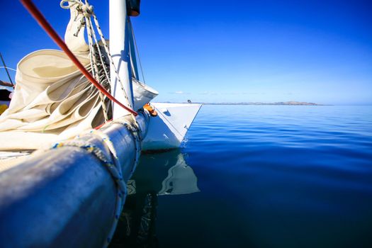 View of the bow of a sailing boat in calm blue sea with red rope riggings and white mast.