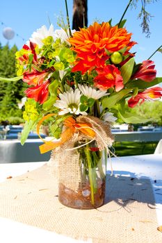 White, orange, and green wedding flower arrangements with burlap as the centerpieces for table settings at an outdoor reception.