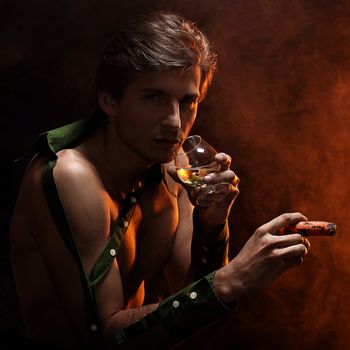 Artistic picture of handsome man with naked torso smoking cigar and drinking wine