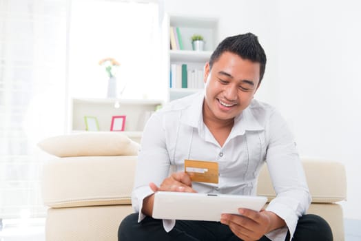 Happy Southeast Asian man online shopping, hands holding credit card and computer tab sitting at home.