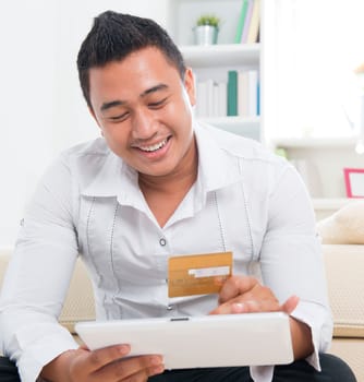 Southeast Asian man online shopping, hands holding credit card and computer tab sitting at home.