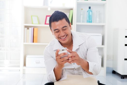 Cheerful Asian male using smartphone at home, looking on screen.