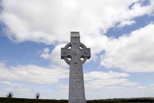 an old celtic cross in an irish graveyard with blue cloudy sky and countryside