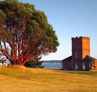 A photograph of an old military building near a waterway.  The large tree in the picture is a Pacific Madrone tree.  The Pacific Madrone (Latin Name: Arbutus menziesii) is a species of tree found on the west coast of North America.  It is known for its distinctive red peeling bark.