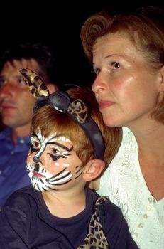 boy and mother are watching a performance, boy has a cat makeup and looks very interrested