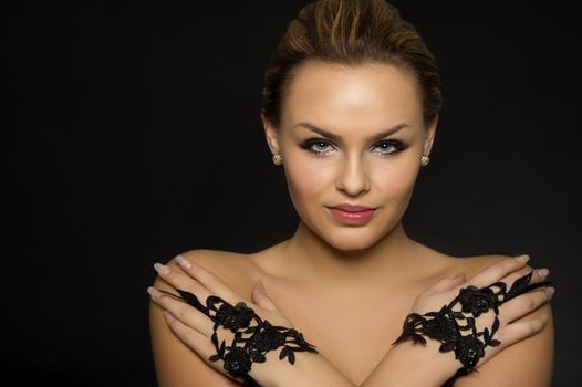 Portrait of a glamorous woman wearing elegant black lace gloves with her hands crossed over her bare shoulders looking at the camera with an enigmatic smile on a dark background with copyspace