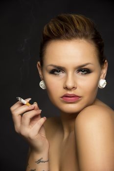 Beautiful seductive woman with bare shoulders and a sultry expression posing against a dark background holding a cigarette between her fingers smoking