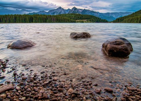 Three rocks in a ripply lake with forest and mountain peaks in the distance.