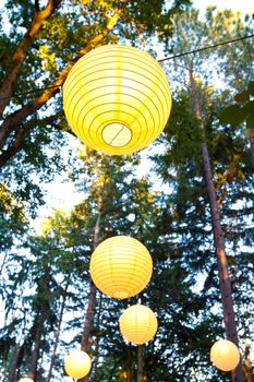 Yellow wedding decorations hang from the trees at a wedding ceremony and reception outdoors in Oregon. These yellow chinese lanterns or japanese lantersn are well lit.