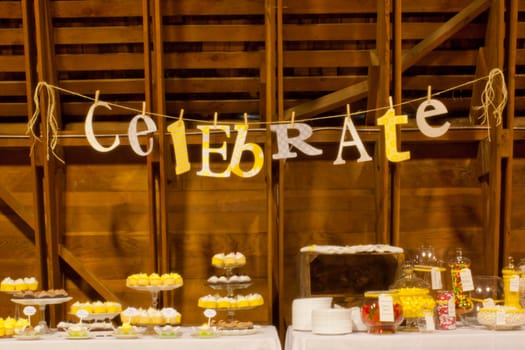 The word celebrate is spelled out on some hung twine in a barn at a wedding reception. Colors chosen use some variety of prints with yellow, white and gray.